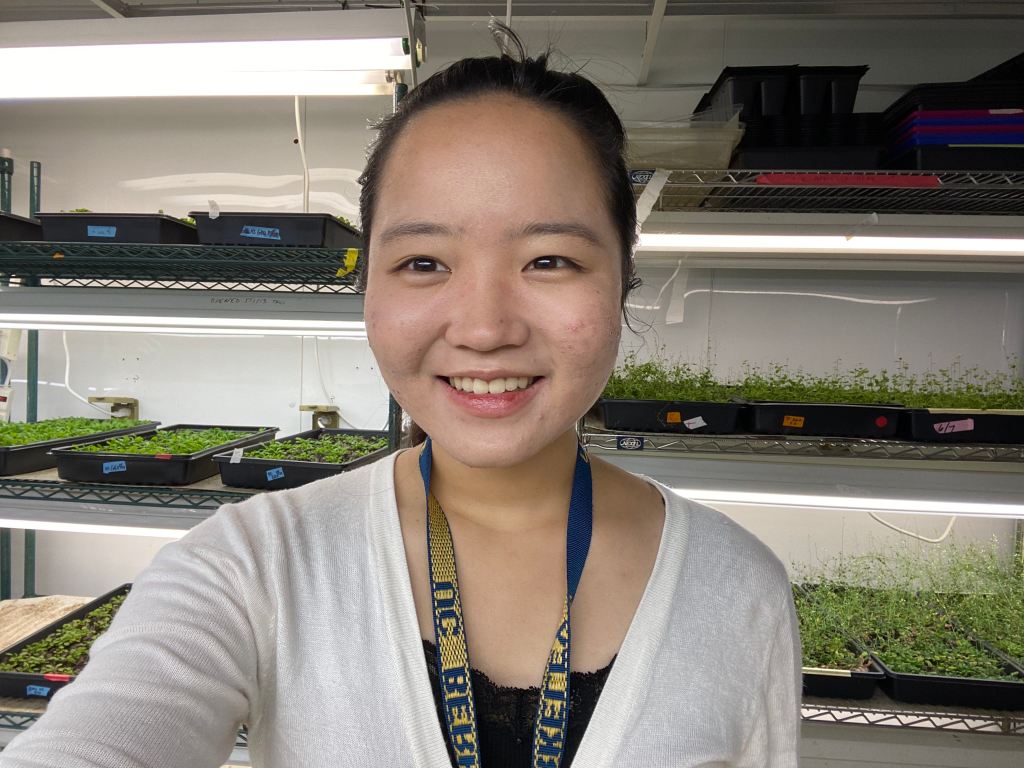Ashleigh Takemoto selfie style portrait wearing a UC Berkeley lanyard, smiling, in a growth chamber in front of shelving units and grow lights stacked with flats of Arabidopsis plants.