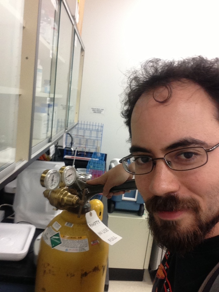 Dr. Benjamin Goldman-Huertas selfie portrait in a lab holding a wrench tightening a regulator onto a gas cylinder.