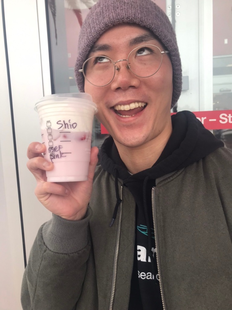 Hiro Suzuki posing in a knit cap looking skywards, showing off a pink and white drink in his right hand.