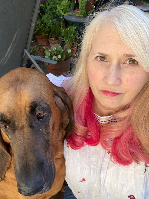 Susan Bernstein posed in front of a shelf of succulents with her dog, Marple, with pink dyed hair tips, wearing a white embroidered top.