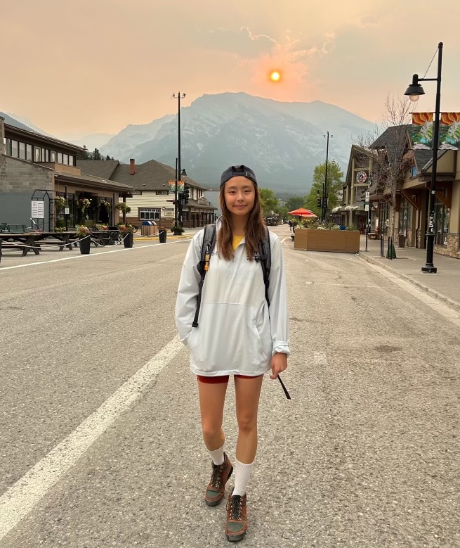 Josephine Tamsil standing near the sidewalk of a small street with a backpack and hicking boots in front of a large snow covered mountain with the setting sun behind her.