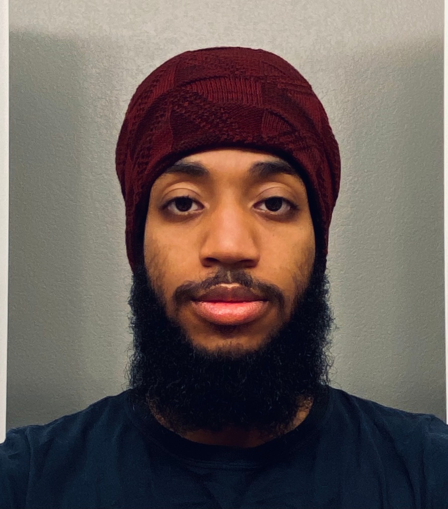 Roem Araujo headshot portrait wearing a red knit cap, looking forward in front of a blank wall.